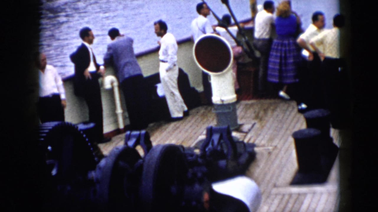 Adults stand on a wooden boat deck near a railing and deck vent as water passes beside them.