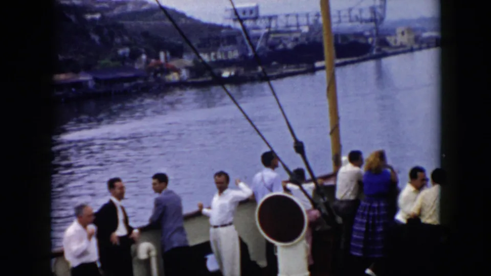 1956 Cuba travel footage frame with passengers on a boat deck, rigging overhead, and a hilly waterfront visible across the water.