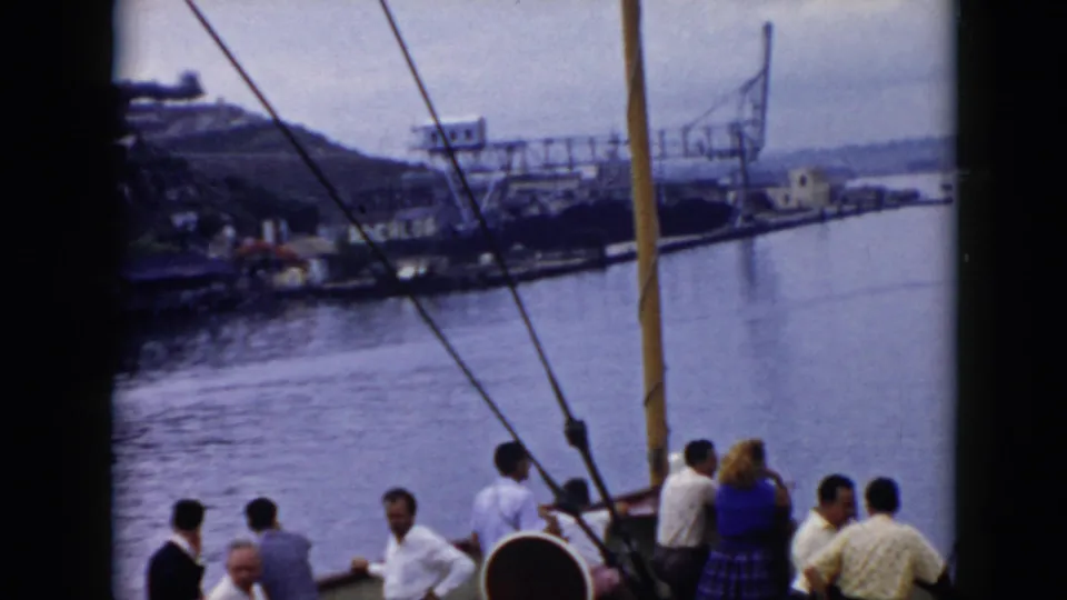 Adults aboard a boat in 1956 Cuba stand near the railing as the camera looks past rigging toward coastal water and distant structures.