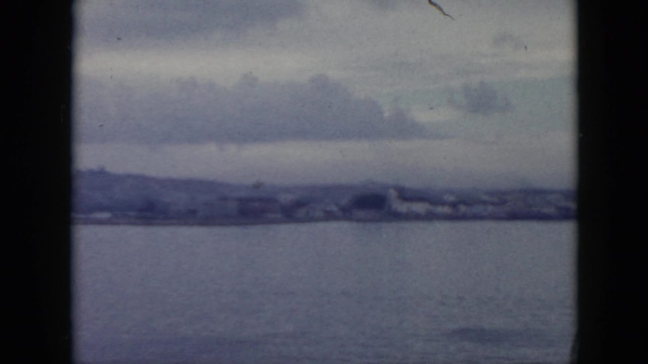 1956 Cuba home-movie view across water toward a hazy shoreline with low buildings, hills, and overcast clouds.