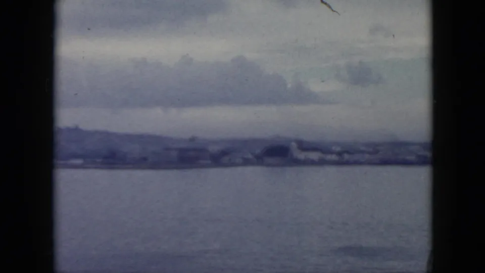 1956 Cuba home-movie view across water toward a hazy shoreline with low buildings, hills, and overcast clouds.