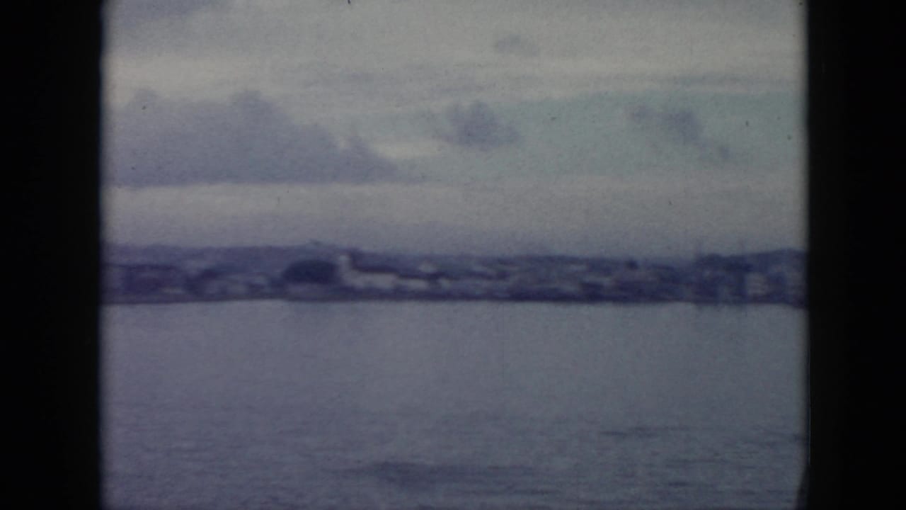Blue-tinted 8mm frame looking across water toward a Cuban shoreline with low structures and distant hills.