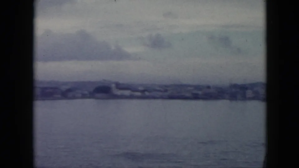 Blue-tinted 8mm frame looking across water toward a Cuban shoreline with low structures and distant hills.