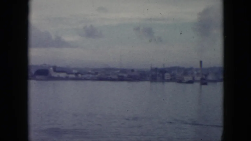 1956 Cuba waterfront view with calm water, distant industrial-looking buildings, a smokestack, and hills beyond.