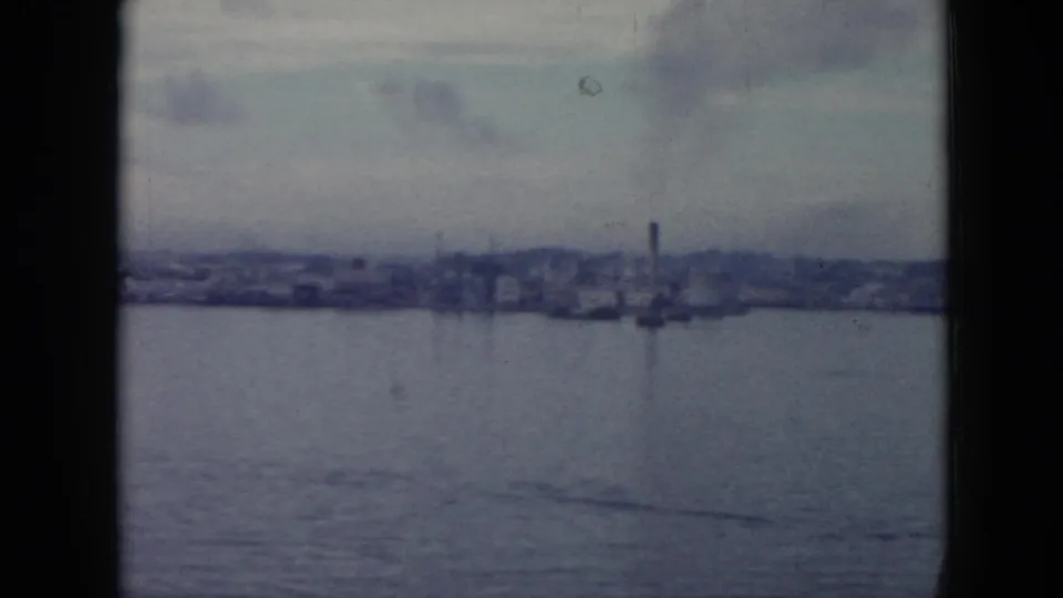 Home-movie frame from Cuba in 1956 showing a hazy waterfront with a smokestack, low buildings, and calm water.