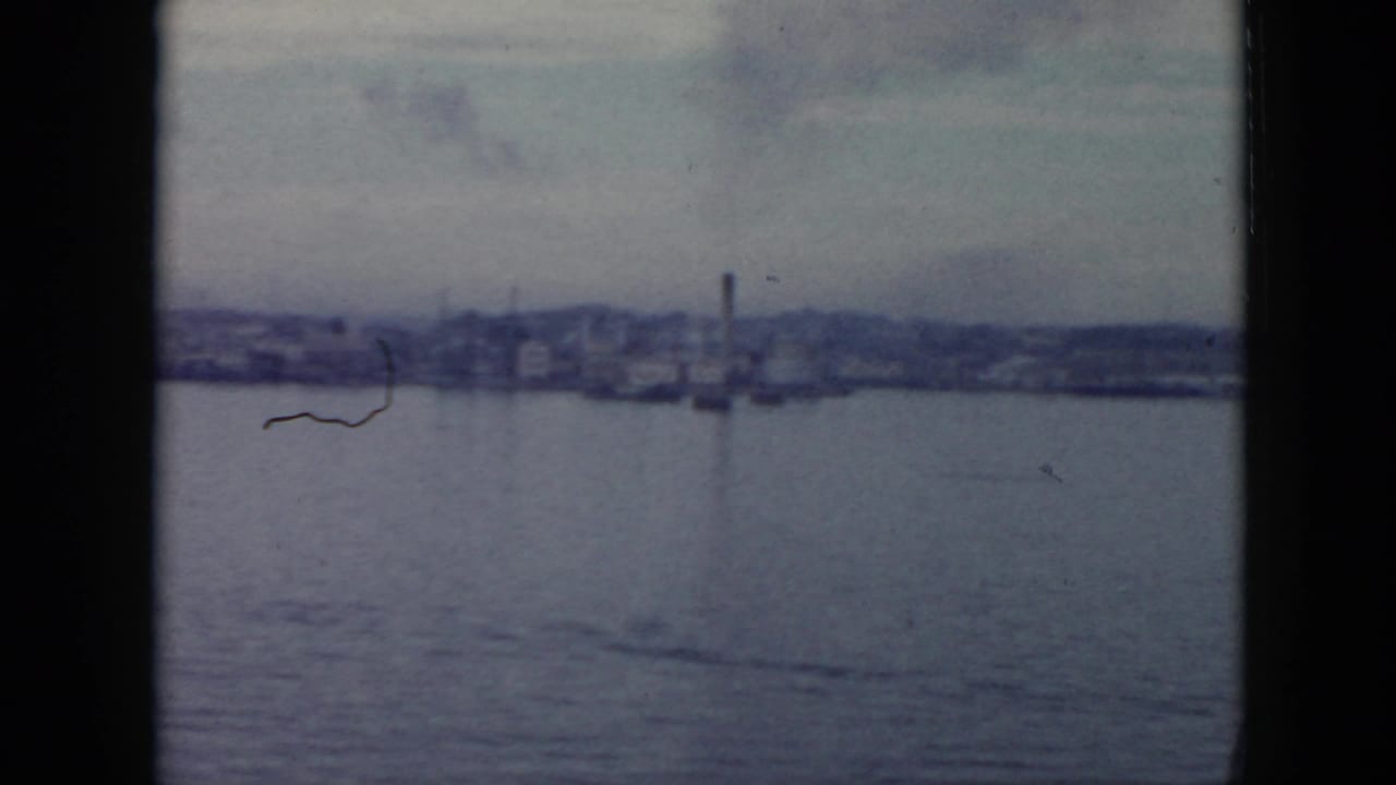 1956 Cuba 8mm frame showing a distant waterfront with low industrial buildings, a smokestack, and calm water in the foreground.