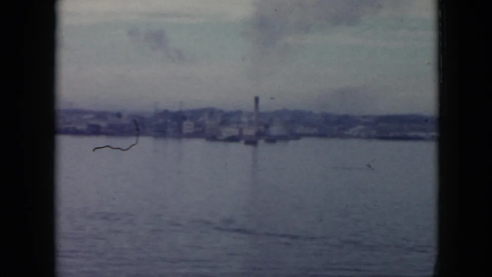 1956 Cuba 8mm frame showing a distant waterfront with low industrial buildings, a smokestack, and calm water in the foreground.