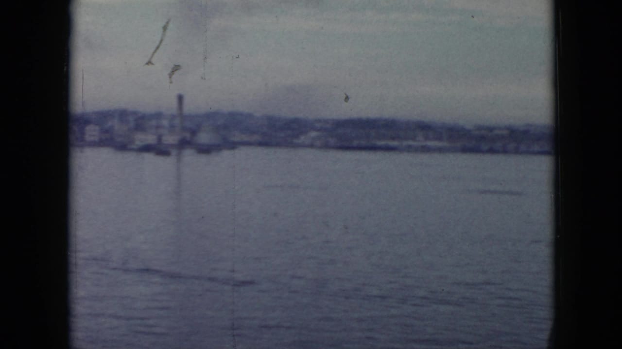 1956 Cuba waterfront view from a boat, with broad water, hazy shoreline buildings, and a vertical smokestack in the distance.