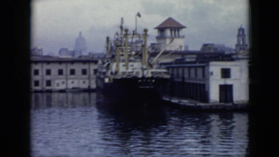 1956 Cuba dockside frame showing a large moored ship beside waterfront buildings, with a small tower and hazy city forms beyond.