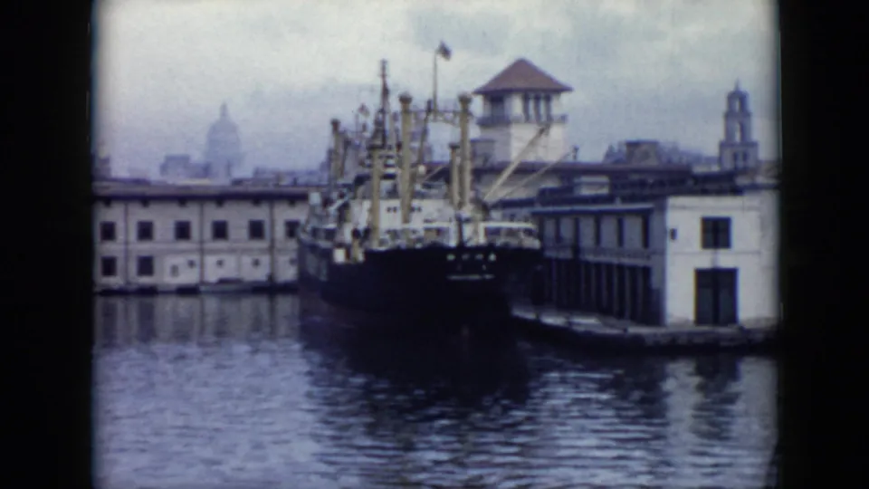 1956 Cuba harbor view with a large cargo ship at a quay, low waterfront buildings, calm water, and a distant dome through haze.