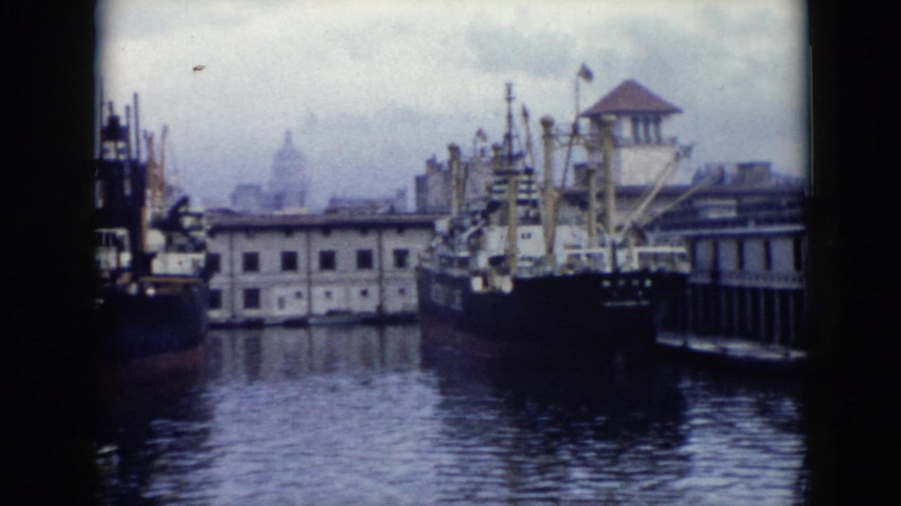 Docked ship and quay buildings seen across harbor water with a hazy city skyline behind.