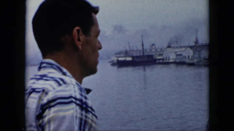 Adult man in a striped shirt stands near the Cuba harbor, seen in profile with docked vessels across the water.