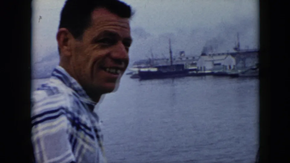 Adult man smiles toward the camera beside a 1956 Cuba harbor view with ships and waterfront buildings behind him.