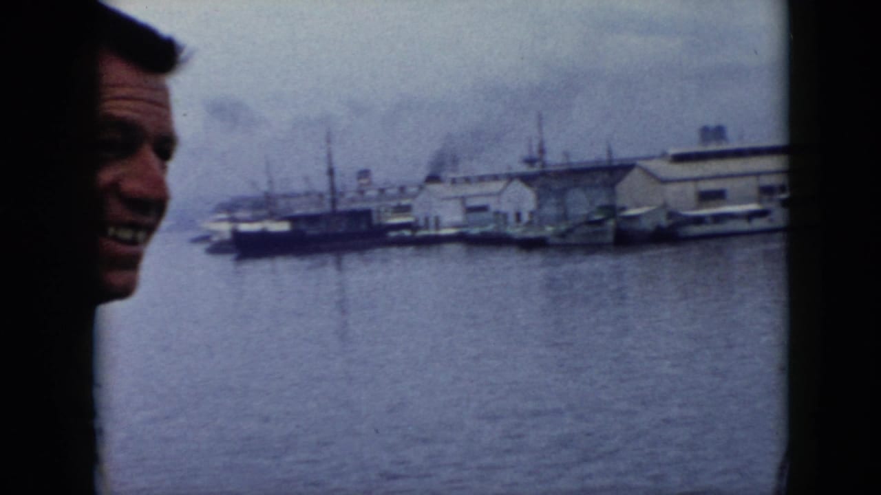 In Cuba 1956 home-movie footage, a smiling adult man appears beside a boat-window view of harbor water, docked boats, and industrial waterfront buildings.