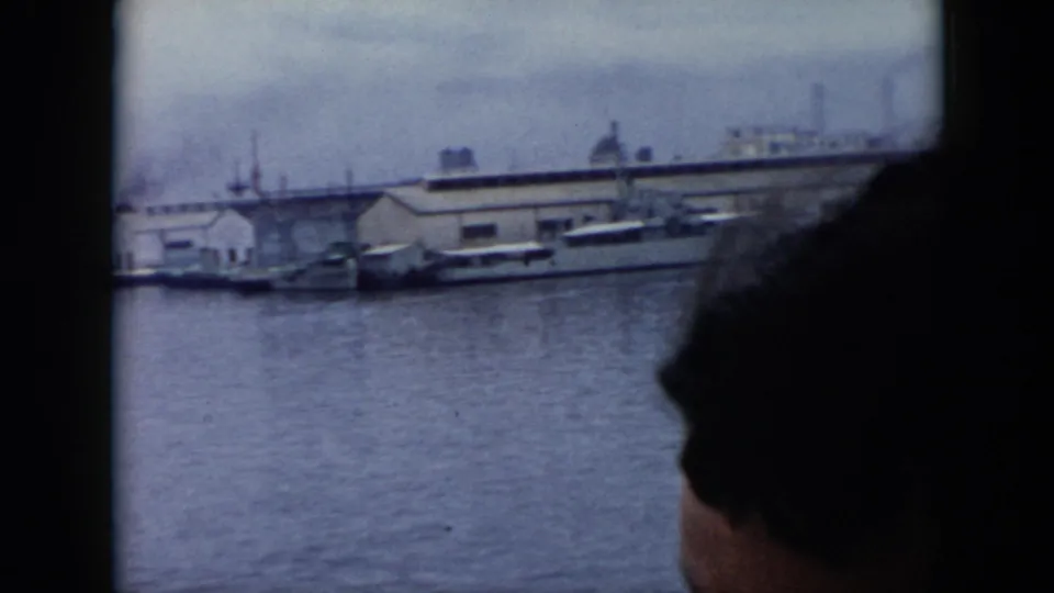 In Cuba 1956 footage, an adult woman is seen in profile from a boat as harbor water, docked vessels, and industrial buildings pass outside.