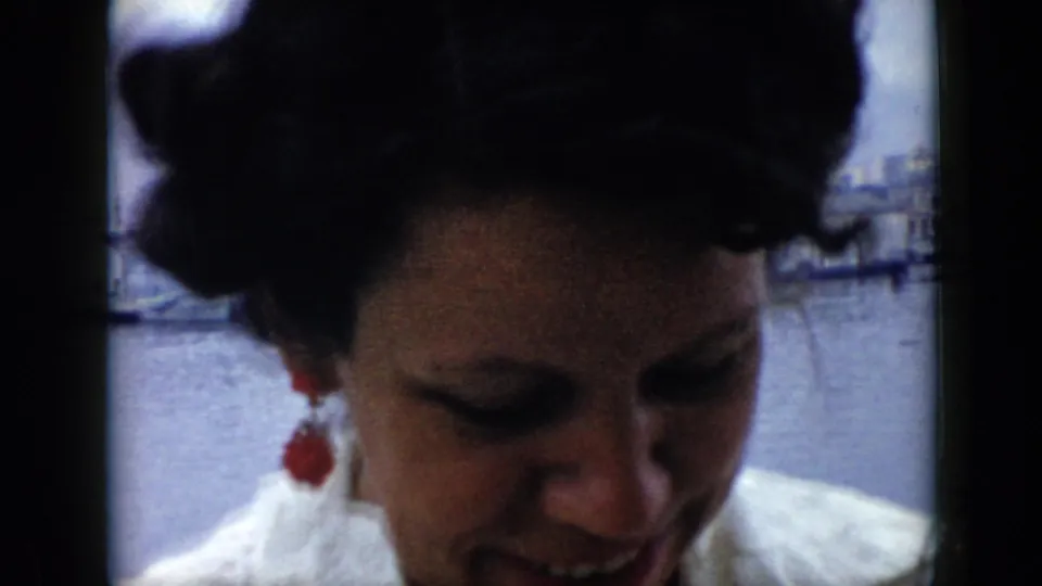 In Cuba 1956 home-movie footage, a woman with dark hair and red earrings smiles on a boat with harbor water and waterfront structures behind her.