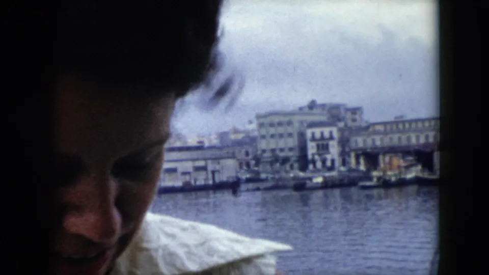 In Cuba 1956 footage, the woman appears partly in shadow on the boat as docked vessels and waterfront buildings line the harbor behind her.