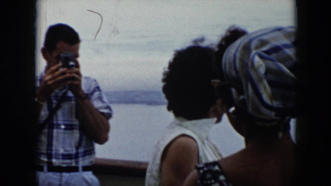 Man in plaid shirt photographs from a ferry while women look toward the harbor.