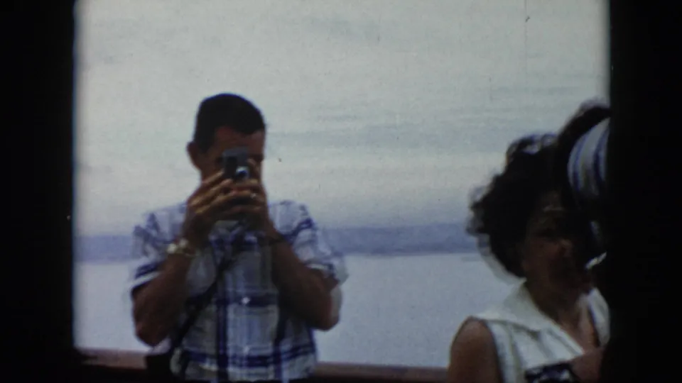 1956 Cuba ferry footage showing a man aiming a camera with women nearby and hazy water in the background.