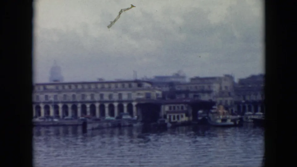 1956 Cuba harbor frame with arched waterfront architecture, boats at shore, cloudy sky, and visible 8mm wear.