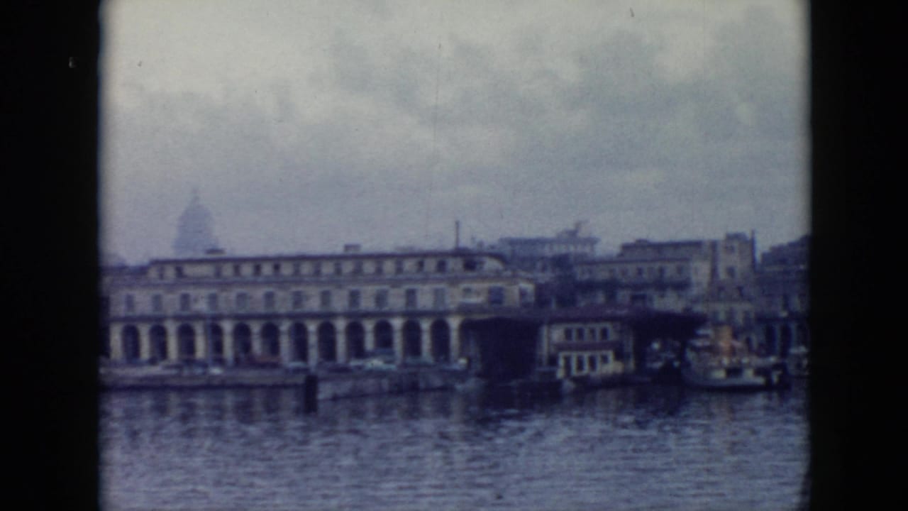 1956 Cuba harbor view with calm water, a long arched waterfront building, dock structures, and overcast sky in soft 8mm color.