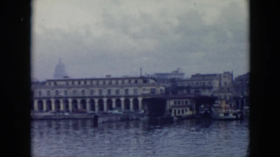 1956 Cuba harbor view with calm water, a long arched waterfront building, dock structures, and overcast sky in soft 8mm color.