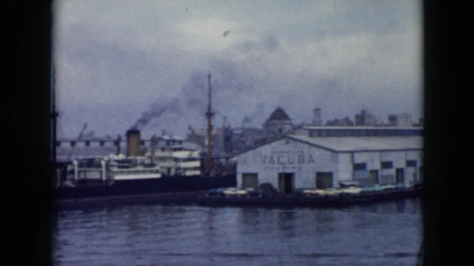1956 Cuba port scene showing a docked vessel, white VACUBA warehouse, cranes, smoke, and calm water in a faded 8mm frame.