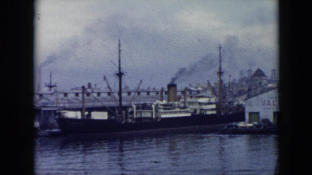 1956 Cuba harbor frame showing a dark-hulled vessel at the waterfront, masts, dock structures, and smoke drifting over the skyline.