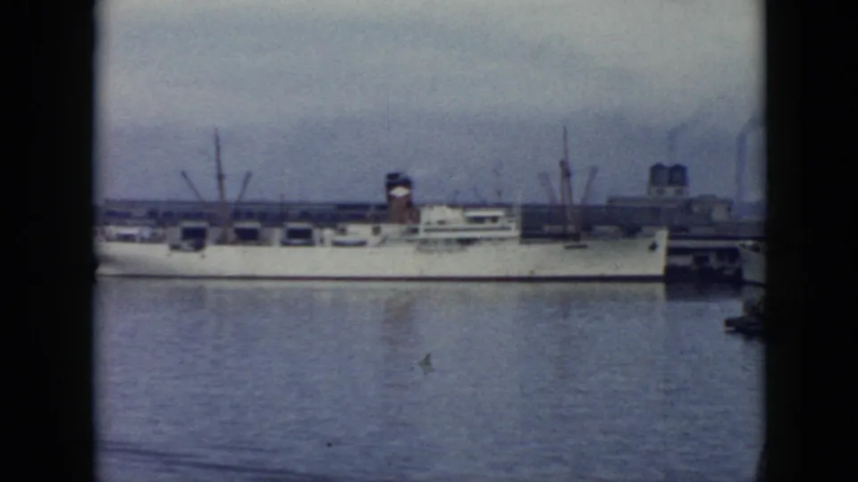 1956 Cuba harbor frame with a long white vessel seen broadside across the water, with dock cranes and industrial structures in the distance.