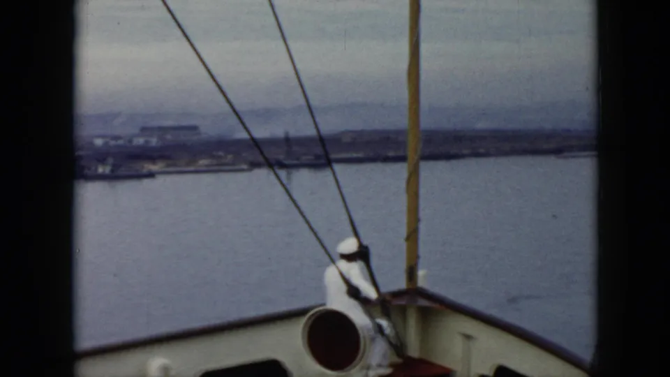 Shipboard 1956 Cuba view with a sailor in white near the helm, rigging lines, railings, harbor water, and distant coastal buildings.