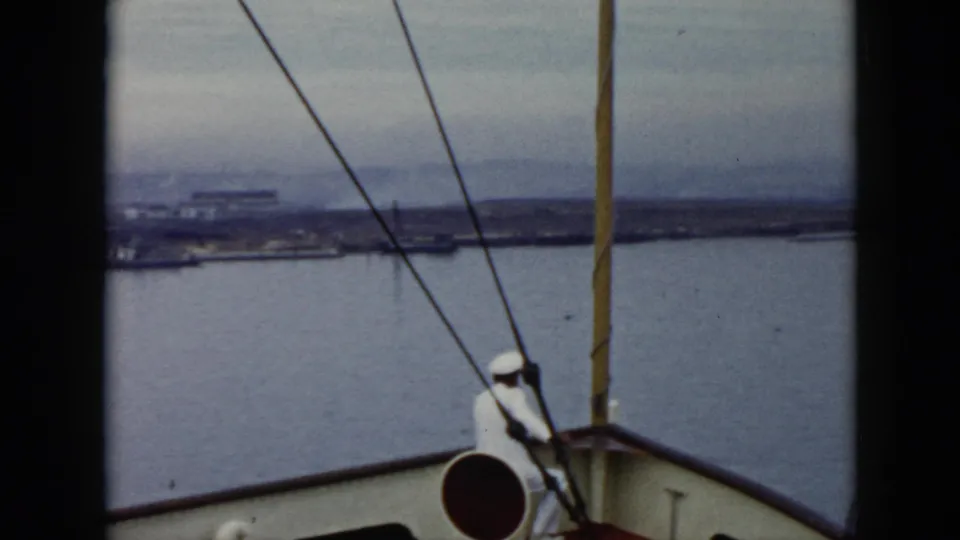 1956 Cuba 8mm shipboard frame looking past a uniformed sailor, rigging, and railings toward harbor water and a low distant shoreline.