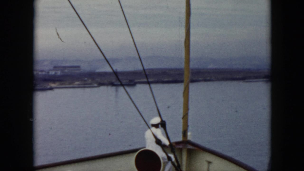 1956 Cuba 8mm frame showing a white-uniformed sailor near the bow of a large boat, with rigging, calm water, and a distant shoreline under a gray sky.