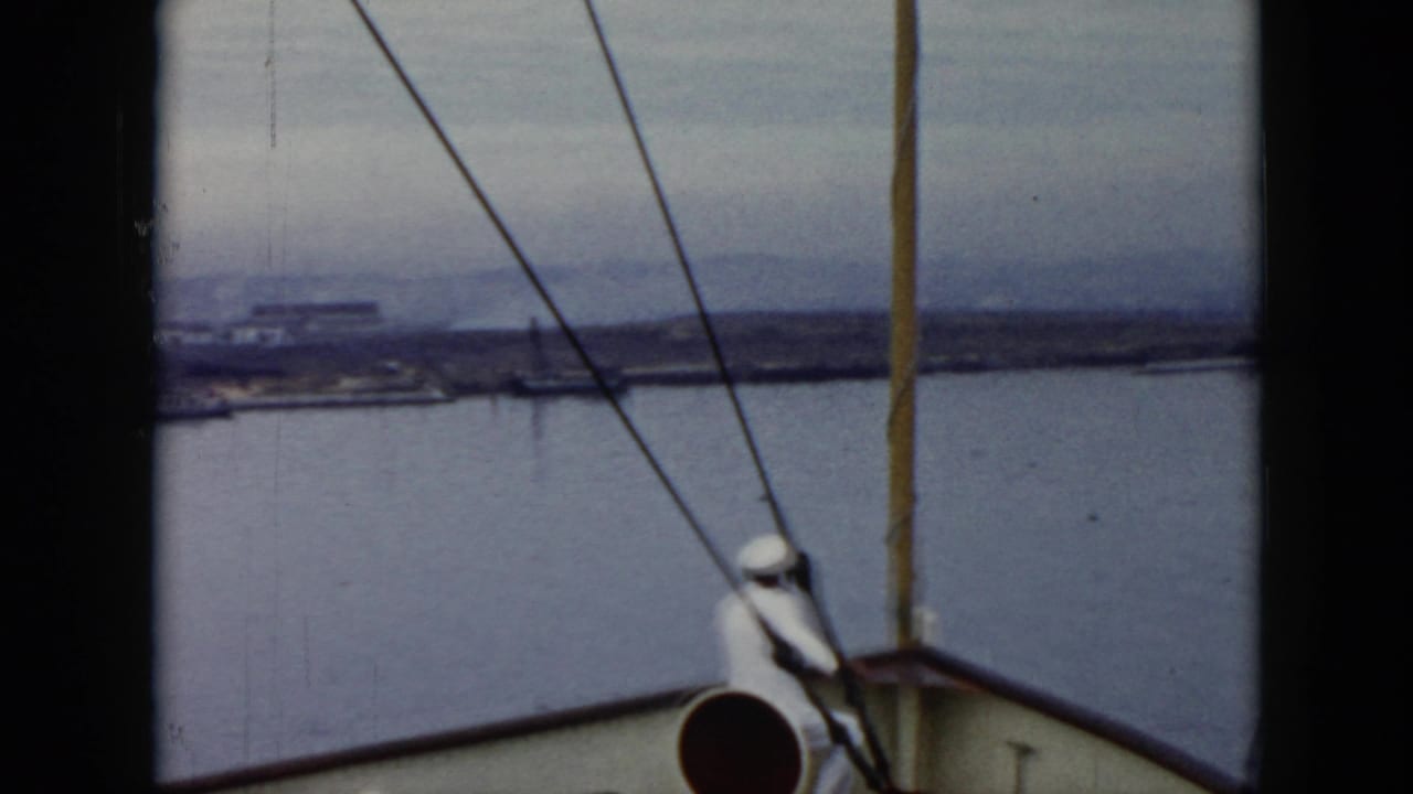Deck-level 1956 Cuba 8mm view with a sailor at the bow, diagonal rigging lines, open water, and low shoreline in the distance.