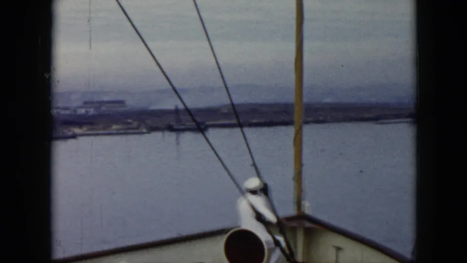 Deck-level 1956 Cuba 8mm view with a sailor at the bow, diagonal rigging lines, open water, and low shoreline in the distance.