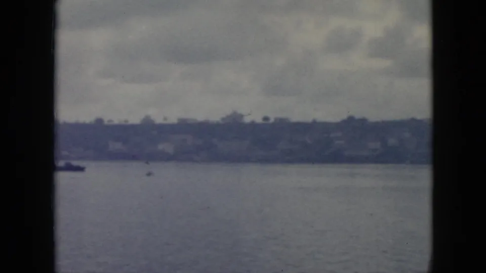 1956 Cuba 8mm shoreline view from a moving boat, showing calm water, low hills, faint buildings, and cloudy sky with no close foreground figure.