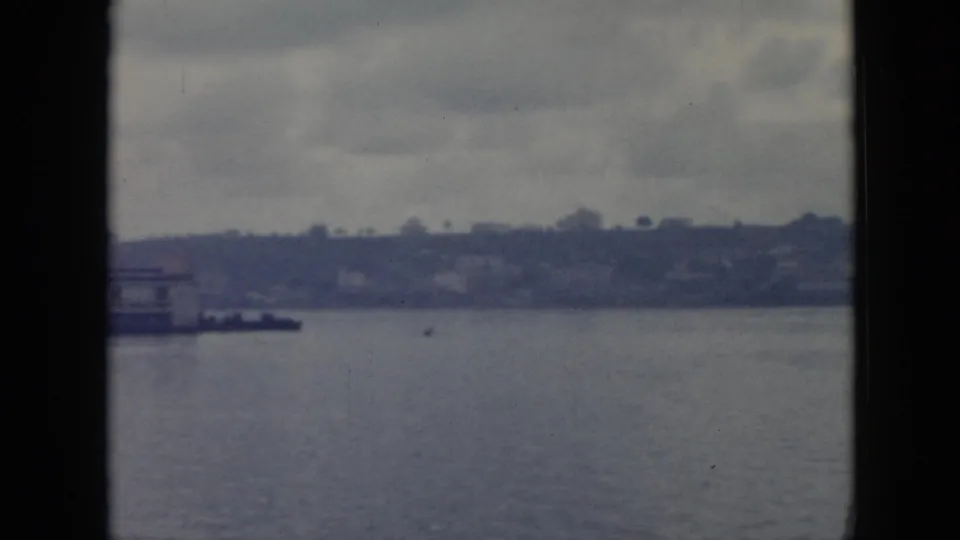 Deck-shot 1956 Cuba 8mm frame looking across water toward a distant shoreline with low structures, hills, and heavy cloud cover.