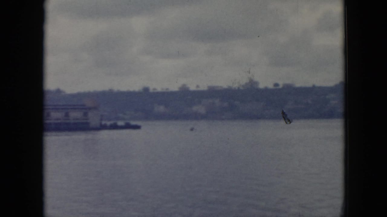 1956 Cuba 8mm frame showing calm water, a distant pier, low buildings, and a soft tree-lined shoreline under gray sky.
