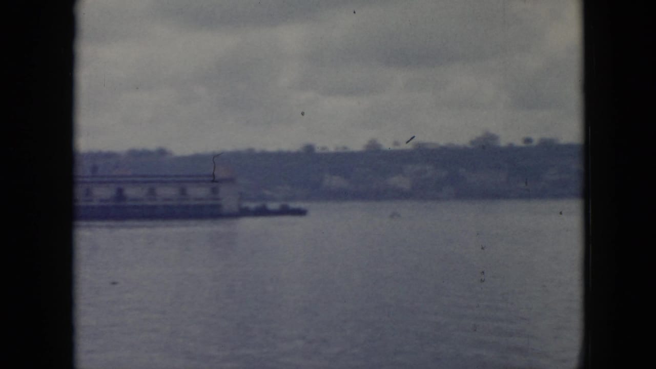 1956 Cuba 8mm view of a quiet waterfront with a low pier, shoreline trees, and calm water in soft blue-gray tones.