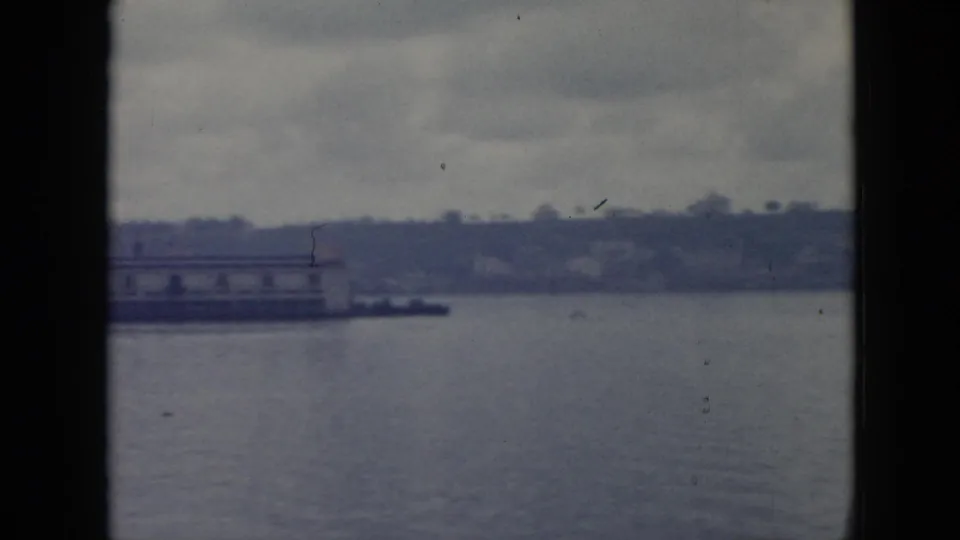 1956 Cuba 8mm view of a quiet waterfront with a low pier, shoreline trees, and calm water in soft blue-gray tones.