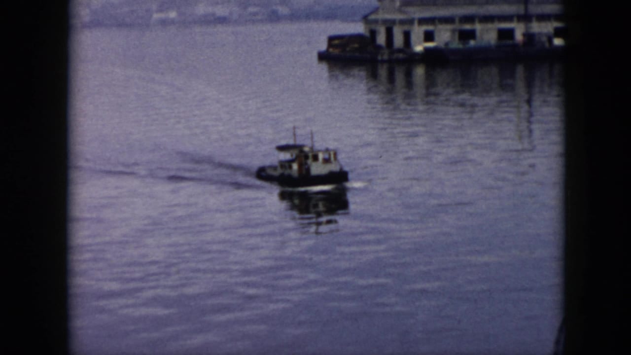 A small tugboat moves across calm harbor water in 1956 Cuba, with dockside buildings and a hazy shoreline behind it.