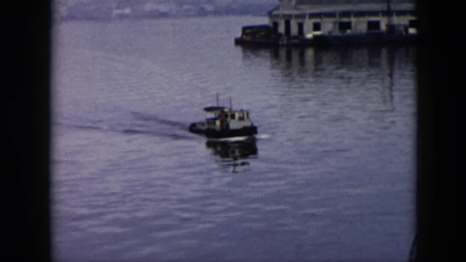A small tugboat moves across calm harbor water in 1956 Cuba, with dockside buildings and a hazy shoreline behind it.