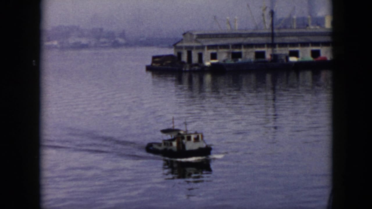 The tugboat continues through calm Cuban harbor water, passing a long dock structure with industrial forms in the background.