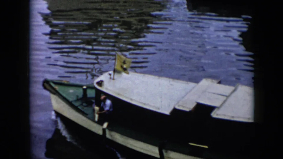 1956 Cuba 8mm frame showing a small covered motorboat moving across harbor water, with a man in a light shirt near the controls and a yellow flag at the bow.