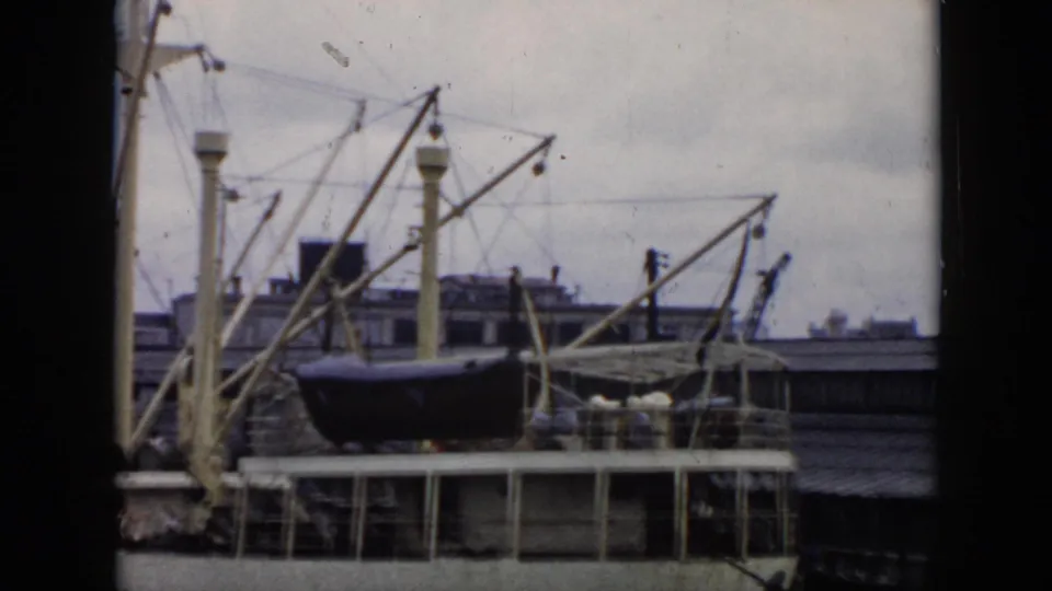 1956 Cuba 8mm frame with a passenger ship, lifeboat, multiple crane arms, pier railings, and soft cloudy background.