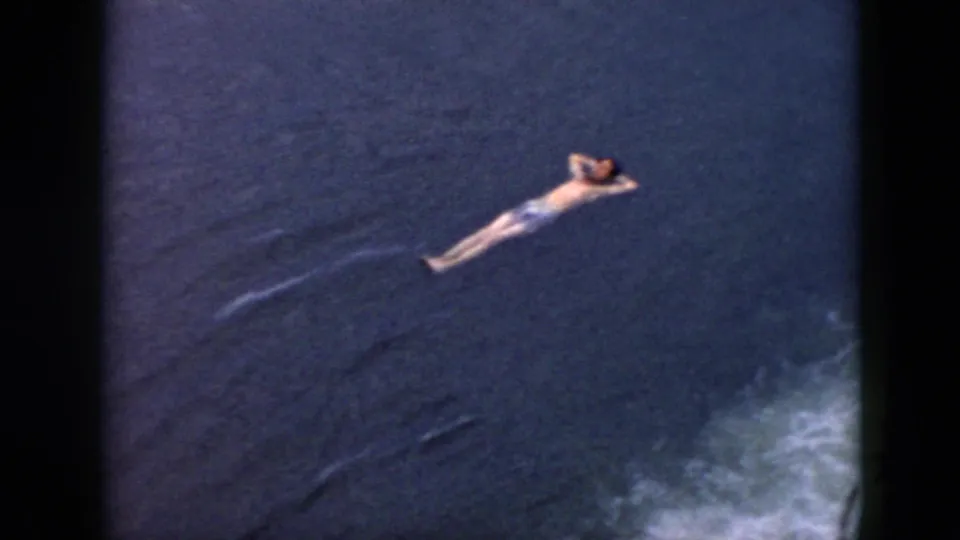 1956 Cuba home-movie frame showing a man swimming or floating on his back across dark lake water.