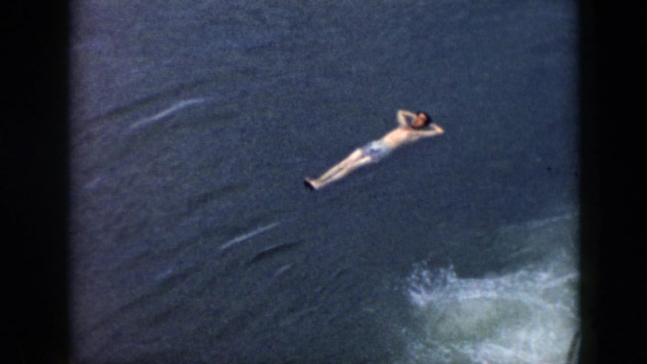 A man floats with hands behind his head in dark water, with white wake visible nearby in this 1956 Cuba home-movie frame.