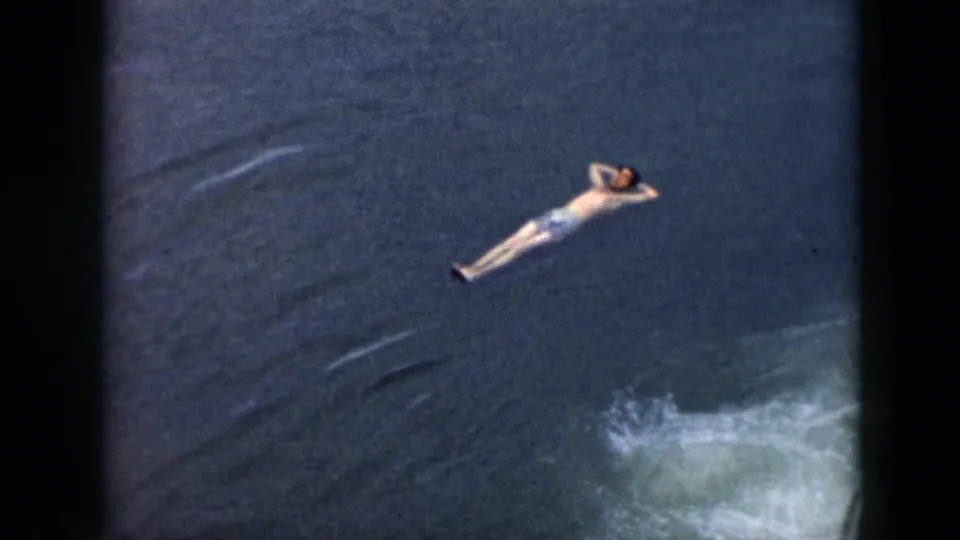 A man floats with hands behind his head in dark water, with white wake visible nearby in this 1956 Cuba home-movie frame.