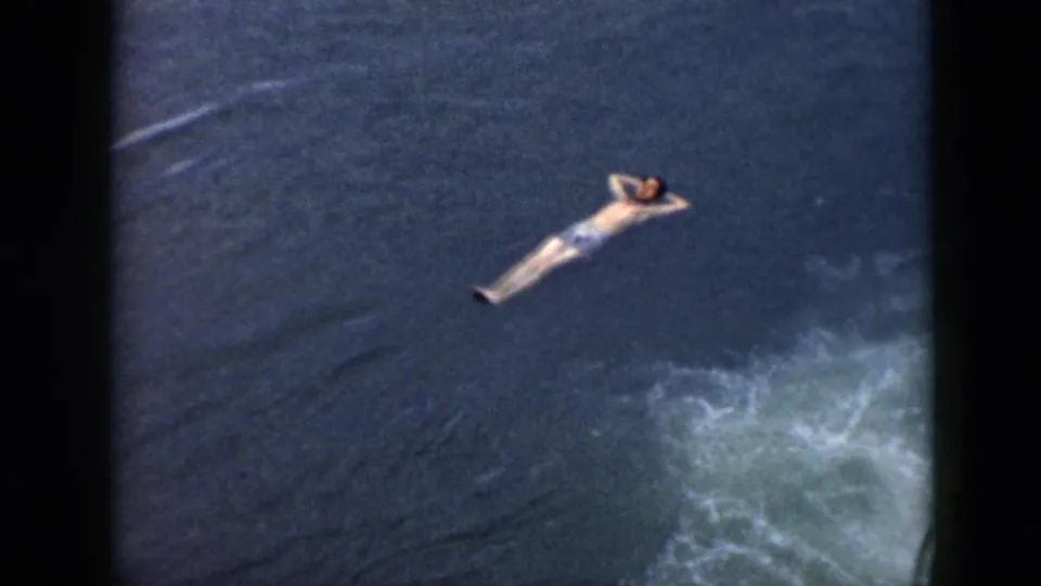 1956 Cuba home-movie frame of a swimmer floating on dark water, with foamy wake entering the lower right of the frame.