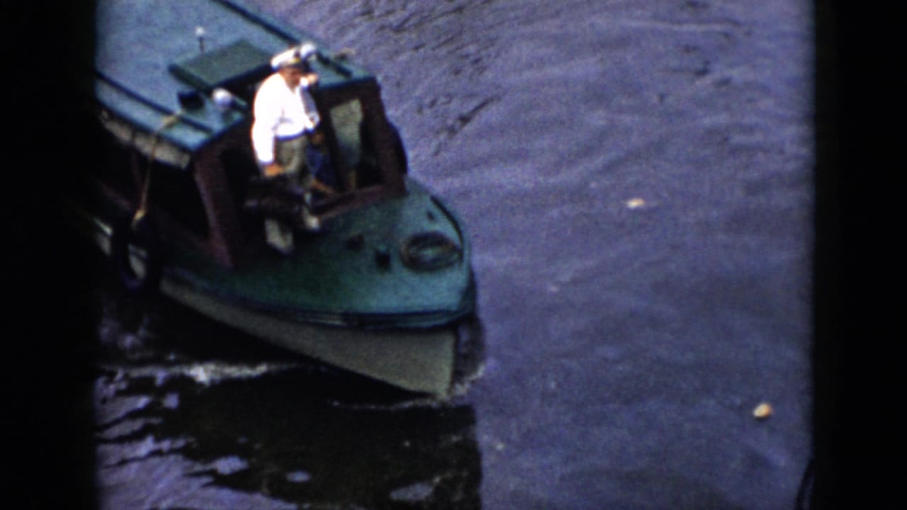 Man in a white sailor cap and shirt stands on the bow of a green cabin motorboat.
