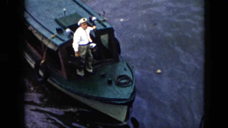 Close 1956 Cuba home-movie view of a man in a white sailor cap on the bow of a green cabin motorboat, with water surrounding the boat.